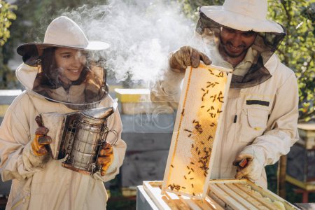 Téléchargez les photos : Heureuse femme apiculteur tenant fumeur par un homme indien apiculteur examinant le cadre en nid d'abeille au jardin rucher - en image libre de droit