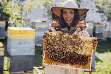 Photo for Happy smiling female Beekeeper in protective suit holding honeybee frame with bees at apiary - Royalty Free Image