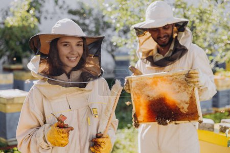 Photo for Couple of happy smiling beekeepers working with beekeeping tools near beehive at bee farm - Royalty Free Image