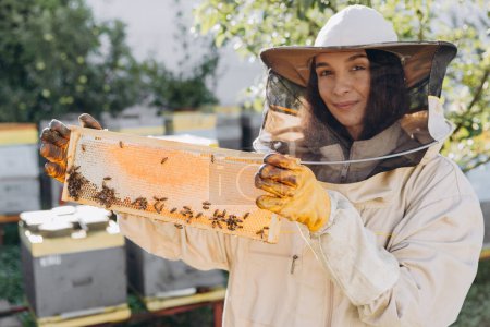 Photo for Happy female Beekeeper in uniform standing in apiary and holding honeybee frame - Royalty Free Image