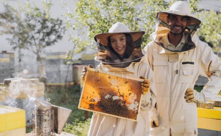 Téléchargez les photos : Couple d'apiculteurs souriants heureux travaillant avec des outils apicoles près de ruche à la ferme apicole - en image libre de droit