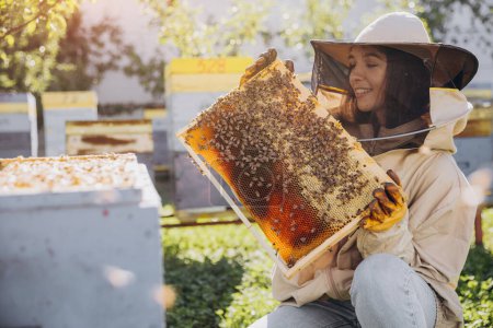 Foto de Feliz hembra apicultora sonriente en traje protector sosteniendo marco de abeja con abejas en el colmenar - Imagen libre de derechos