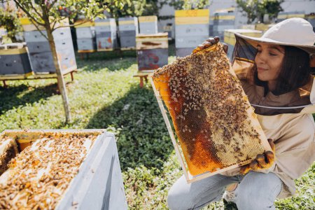 Feliz hembra apicultora sonriente en traje protector sosteniendo marco de abeja con abejas en el colmenar