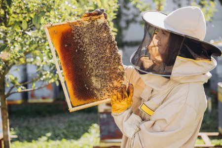 Joyeux souriant femme apiculteur en uniforme debout dans le rucher et tenant cadre abeille