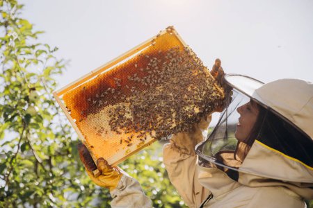 Feliz hembra apicultora sonriente en uniforme de pie en colmenar y con marco de abeja