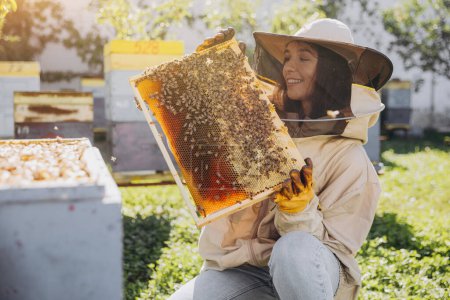 Glücklich lächelnde Imkerin im Schutzanzug hält Bienenrahmen mit Bienen am Bienenstand