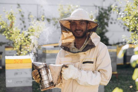 Foto de Feliz sonriente macho indio apicultor fumando abejas con abeja fumador en el colmenar - Imagen libre de derechos