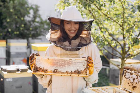 Téléchargez les photos : Heureux apiculteur féminin en uniforme debout dans le rucher et tenant cadre abeille - en image libre de droit