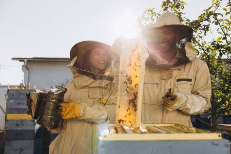 Photo for Happy woman beekeeper holding smoker by indian man apiarist examining honeycomb frame at apiary garden - Royalty Free Image