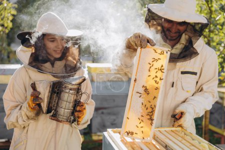 Happy woman beekeeper holding smoker by indian man apiarist examining honeycomb frame at apiary garden