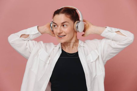 Photo for Modern woman smiling with braces on her teeth and listening to music in headphones on pink background - Royalty Free Image