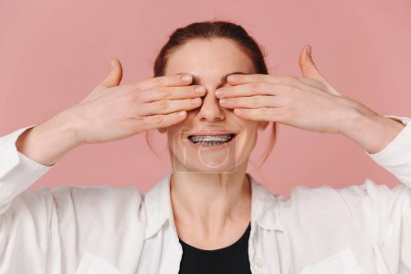 Foto de Mujer feliz moderna sonriendo ampliamente con aparatos ortopédicos y cubriendo los ojos con las manos sobre fondo rosa - Imagen libre de derechos
