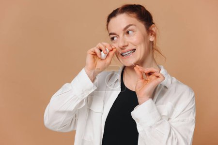 Téléchargez les photos : Portrait de femme mature dans des bretelles tenant du fil dentaire près de ses dents sur fond beige - en image libre de droit