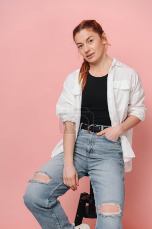 Photo for Modern sporty woman with red hair wearing shirt and jeans posing on pink background - Royalty Free Image