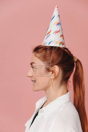 Foto de Retrato de mujer moderna sonriente con tirantes y sombrero de cumpleaños de pie de perfil sobre fondo rosa - Imagen libre de derechos