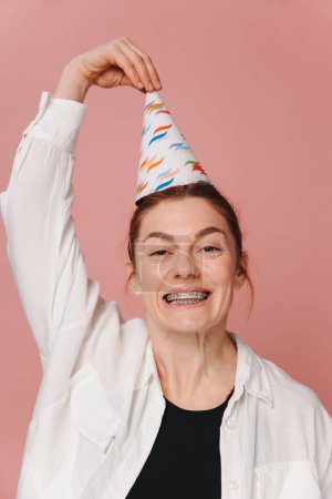 Téléchargez les photos : Portrait de femme moderne souriant avec des bretelles et grimaçant et tenant chapeau d'anniversaire sur fond rose - en image libre de droit