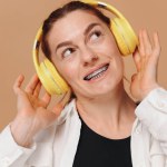 Modern woman smiling with braces on her teeth and listening to music in headphones on a beige background