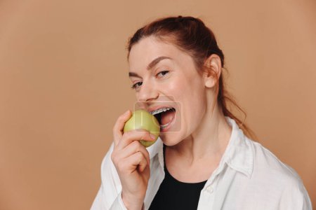 Retrato de mujer madura con tirantes en los dientes comiendo manzana verde sobre fondo beige