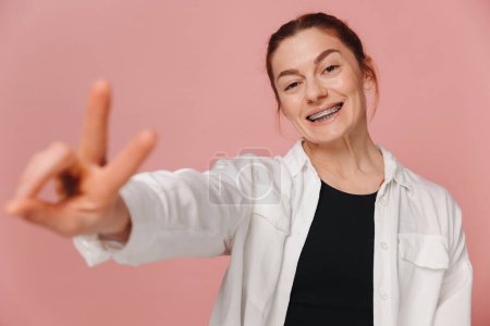 Photo for Modern happy smiling woman with braces showing peace on pink background - Royalty Free Image
