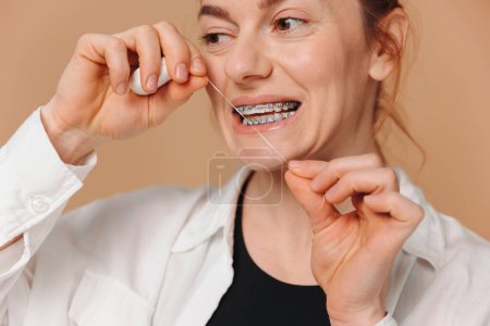 Téléchargez les photos : Portrait en gros plan d'une femme mûre portant du fil dentaire près de ses dents sur fond beige - en image libre de droit