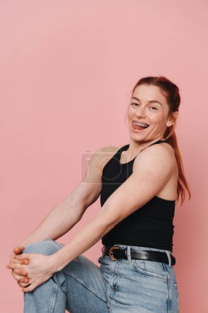 Sportliche, muskulöse Frau in T-Shirt und Jeans, die emotional lacht, mit Hosenträgern auf rosa Hintergrund