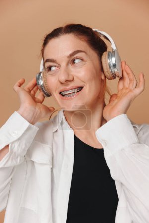 Photo for Modern woman smiling with braces on her teeth and listening to music in headphones on a beige background - Royalty Free Image