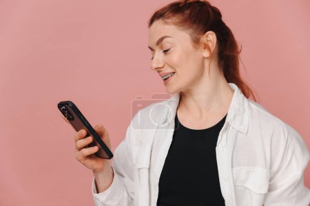 Photo for Modern happy woman smiling widely with braces and talking on smartphone on pink background - Royalty Free Image