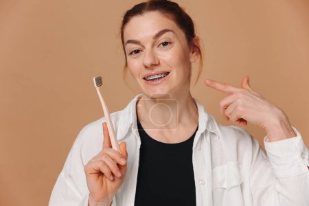 Téléchargez les photos : Heureuse femme mature dans des bretelles tenant une brosse à dents sur un fond beige - en image libre de droit