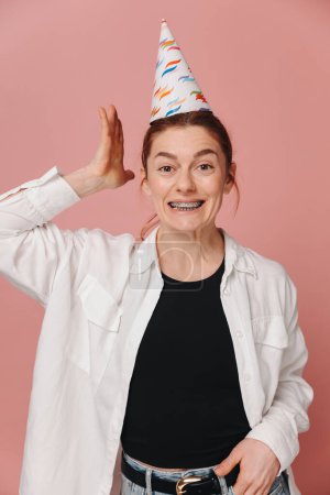 Téléchargez les photos : Portrait de femme moderne souriante avec bretelles et grimacer dans un chapeau d'anniversaire sur fond rose - en image libre de droit