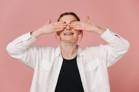 Foto de Mujer feliz moderna sonriendo ampliamente con aparatos ortopédicos y cubriendo los ojos con las manos sobre fondo rosa - Imagen libre de derechos