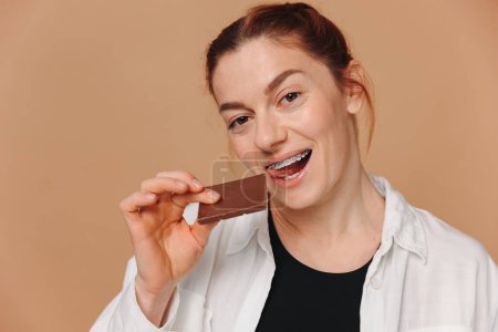 Photo for Portrait of mature woman in braces biting chocolate on a beige background - Royalty Free Image
