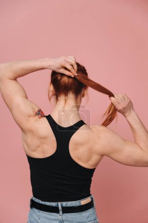 Photo for Sporty muscular woman in t-shirt and jeans showing strong back on pink background - Royalty Free Image
