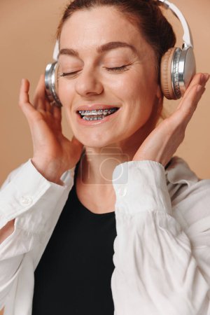Téléchargez les photos : Femme moderne souriant avec des accolades sur ses dents et écoutant de la musique dans les écouteurs sur un fond beige - en image libre de droit
