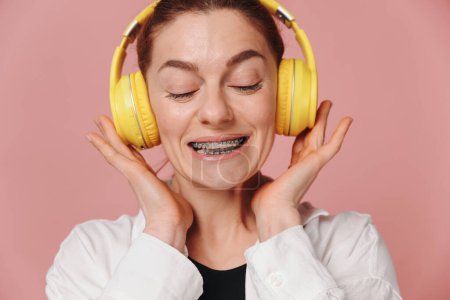 Modern woman smiling with braces on her teeth and listening to music in headphones on pink background