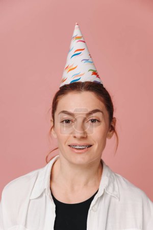Photo for Portrait of smiling modern woman with braces and wearing birthday hat on pink background - Royalty Free Image