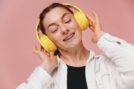 Photo for Modern woman smiling with braces on her teeth and listening to music in headphones on pink background - Royalty Free Image