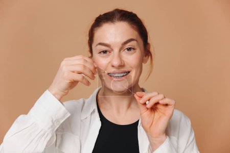 Téléchargez les photos : Portrait de femme mature dans des bretelles tenant du fil dentaire près de ses dents sur fond beige - en image libre de droit
