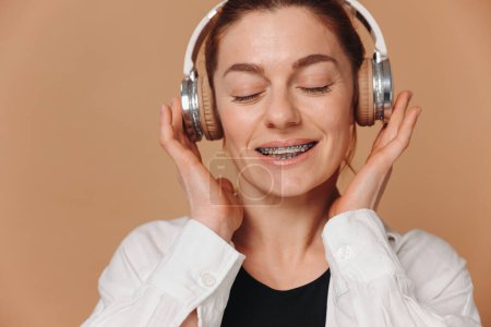 Téléchargez les photos : Femme moderne souriant avec des accolades sur ses dents et écoutant de la musique dans les écouteurs sur un fond beige - en image libre de droit