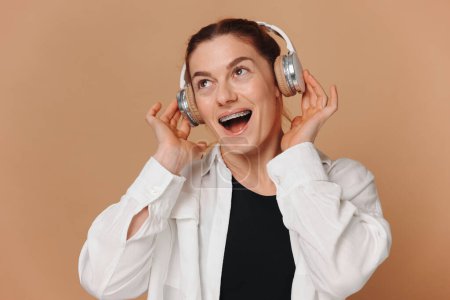 Foto de Mujer moderna sonriendo con frenillos en los dientes y escuchando música en auriculares sobre un fondo beige - Imagen libre de derechos