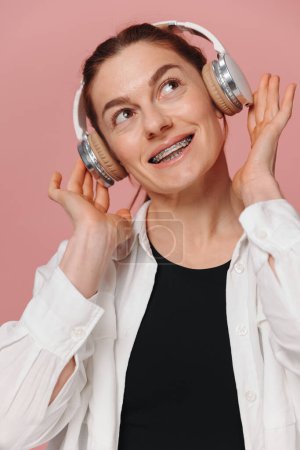 Téléchargez les photos : Femme moderne souriant avec des accolades sur ses dents et écoutant de la musique dans les écouteurs sur fond rose - en image libre de droit