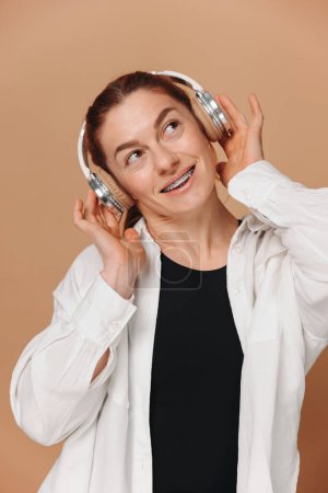 Mujer moderna sonriendo con frenillos en los dientes y escuchando música en auriculares sobre un fondo beige