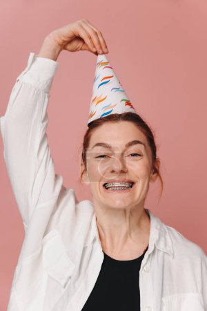 Foto de Retrato de mujer moderna sonriendo con frenos y muecas y sosteniendo sombrero de cumpleaños sobre fondo rosa - Imagen libre de derechos
