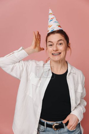 Foto de Retrato de mujer moderna sonriente con frenos y muecas en sombrero de cumpleaños sobre fondo rosa - Imagen libre de derechos