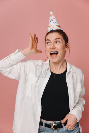 Photo for Portrait of smiling modern woman with braces and grimacing in birthday hat on pink background - Royalty Free Image