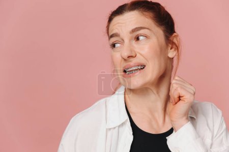 Photo for Portrait of red-haired mature woman suffering from pain due to braces on pink background - Royalty Free Image