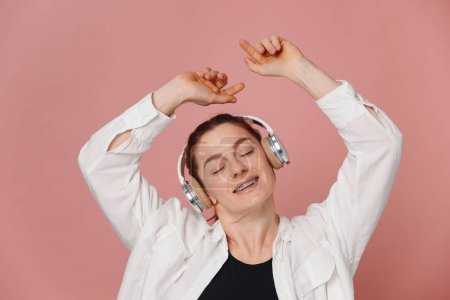 Foto de Mujer moderna sonriendo con aparatos ortopédicos en los dientes y escuchando música en auriculares y bailando sobre fondo rosa - Imagen libre de derechos