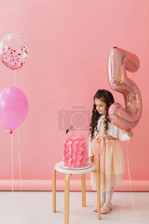 Foto de Adorable niña en un vestido rosa posando junto a un pastel de cumpleaños flamenco y celebrando su quinto cumpleaños sobre un fondo rosa - Imagen libre de derechos