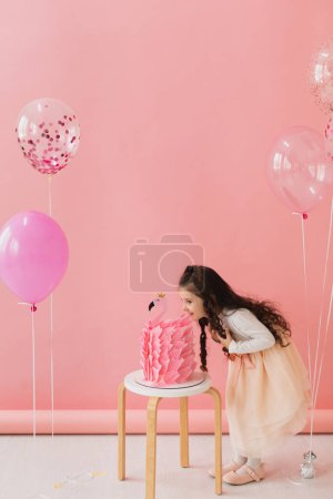 Téléchargez les photos : Adorable petite fille dans une robe rose posant à côté d'un gâteau d'anniversaire flamant rose et célébrant son cinquième anniversaire sur un fond rose - en image libre de droit
