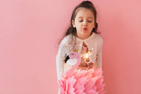 Téléchargez les photos : Adorable petite fille dans une robe rose posant à côté d'un gâteau d'anniversaire flamant rose et célébrant son cinquième anniversaire sur un fond rose - en image libre de droit