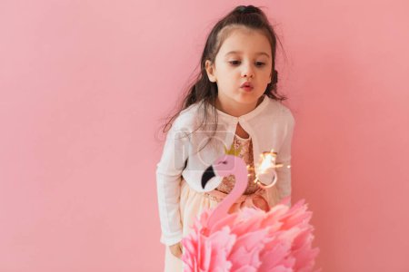 Photo for Adorable little girl in a pink dress posing next to a flamingo birthday cake and celebrating her fifth birthday on a pink background - Royalty Free Image